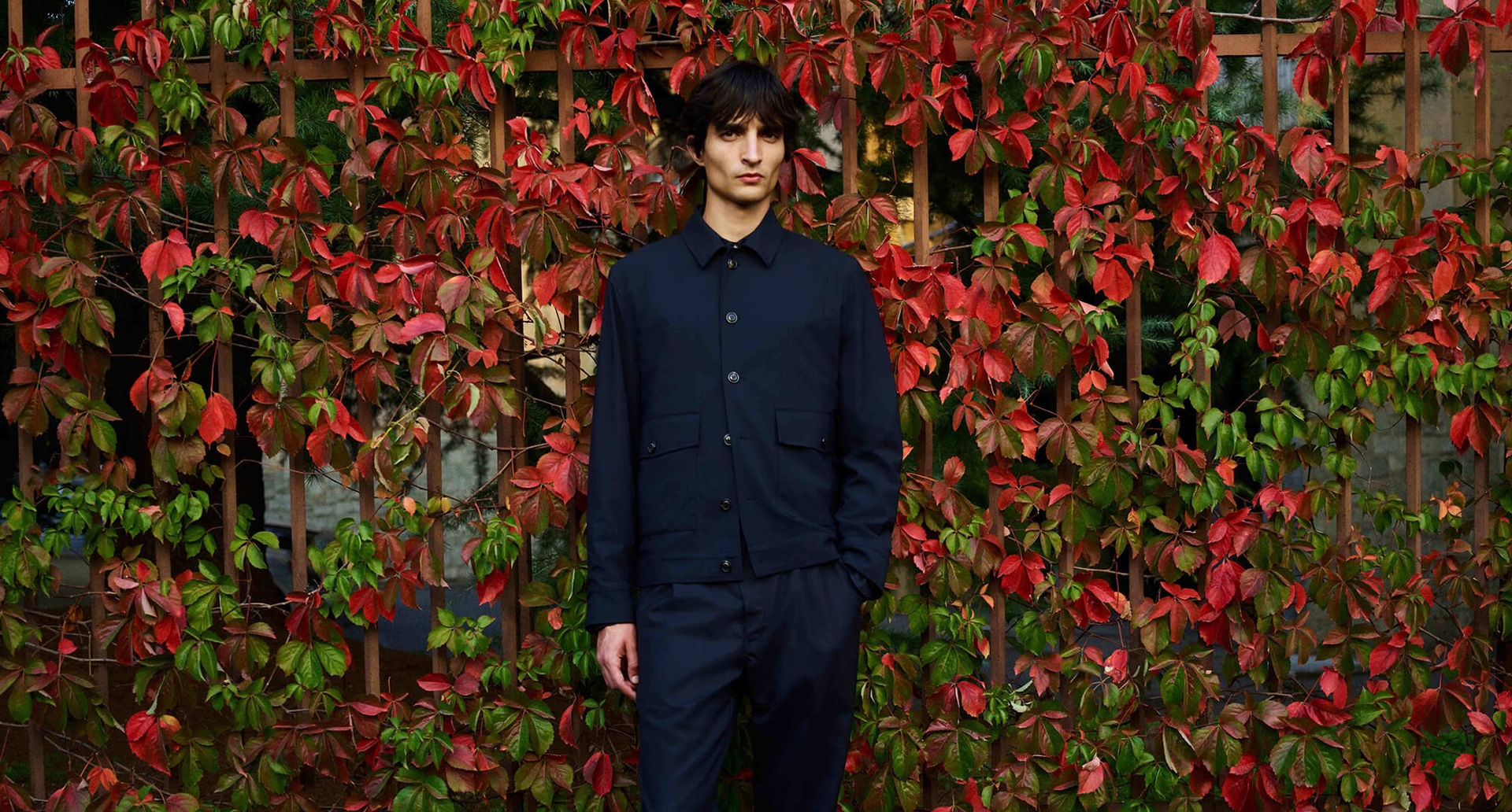 A man wearing a navy Valstar Milano jacket standing in front of red and green leaves